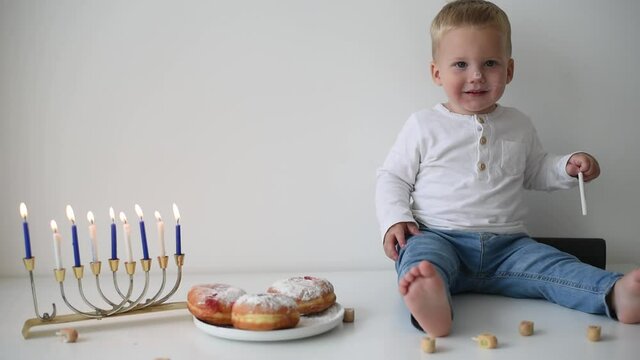 Kid in white t-shirt on white background celebrating Hanukkah. Jewish festival of lights. Child smile and happy. On table donuts and traditional menorah with lighting candles. Holiday in Israel 