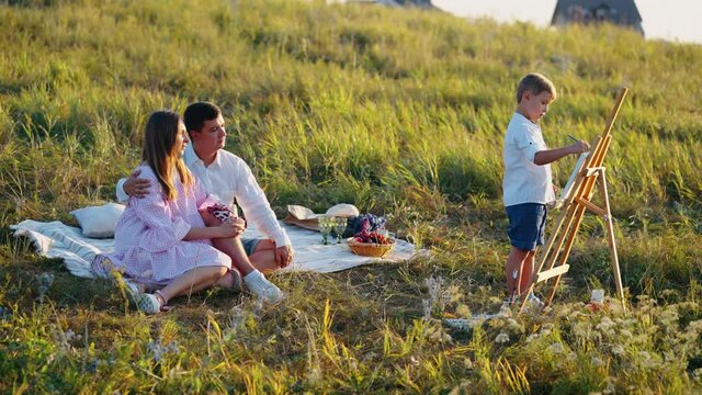 Happy family having picnic in meadow, parents hugging on blanket and watching little son drawing picture on canvas. Enjoying creative hobby in nature. Concept of arts