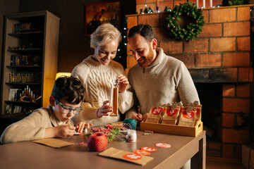 Smiling young family making Christmas advent calendar together sitting at table on background of fireplace in cozy house on xmas Eve, enjoying togetherness preparing for holidays at home.