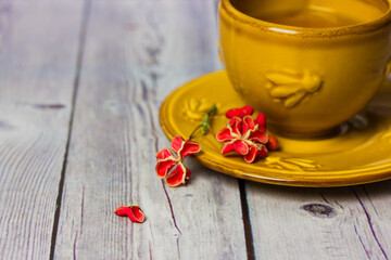 Ceramic yellow cup with black tea stands on saucer on grey wooden table. Spindle shrub, Euonymus europaeus, Pfaffenhutchen red flower petals. Petal in a form of heart. Flat lay still life composition.