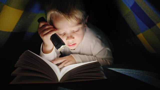 Boy child to make a camp to play imaginatively and reading book by flashlight in living room at home.
