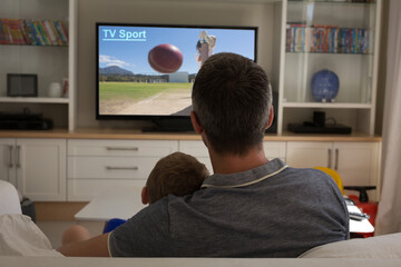 Rear view of father and son sitting at home together watching sport event on tv
