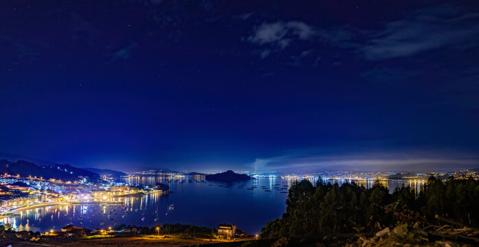 Panor&aacute;mica de pueblos costeros de noche en la R&iacute;a de Pontevedra con playa, embarcadero, barcos,  bateas de mejillones y isla y ciudad de fondo con luces.