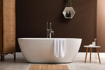 Modern ceramic bathtub with towel near brown wall in room