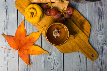 Autumnal still life - bunch of grapes, ripe quince, ginger root lie on wooden cutting board top view. Orange maple leaf on table. Yellow cup with hot aromatic tea drink decorated with flower. 