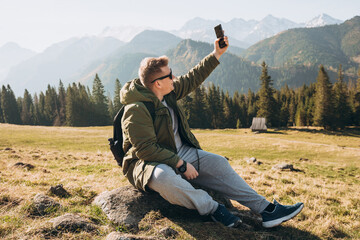 Young man in sunglasses making selfie photo high in the snow mountains enjoying the view. Freedom, happiness, travel and vacations concept, outdoor activities, he wearing a green jacket