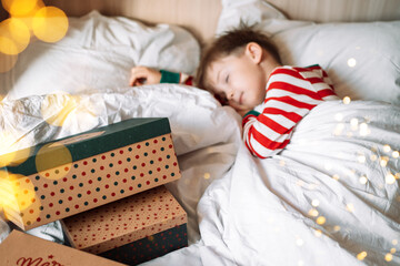 Boy sleeping on white bedding next to christmas gifts