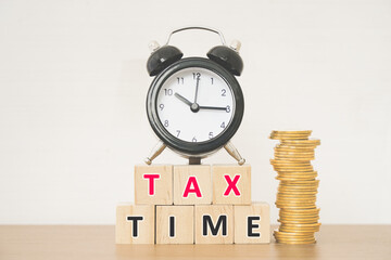 tax time word on wooden block, black analog alarm clock ant stack of coins on wood desk