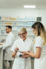 Obraz premium Senior female pharmacist using a tablet while working in a pharmacy with her young colleague