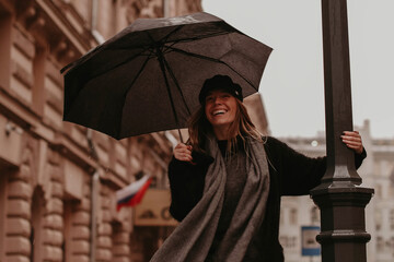 Portrait of a young laughing woman in a cozy black autumn warm coat and a gray scarf walking with an umbrella in the rain. Street casual female fashion © asauriet