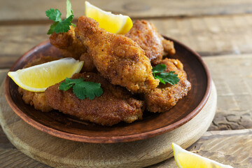 Breaded crispy chicken wings and legs with lemon wedges and cilantro leaves in a ceramic plate on a wooden background. Traditional food concept. Horisontal orientation.