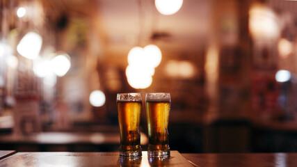 two glasses of cold beer against the background of a blurry empty beer house interior - obrazy, fototapety, plakaty