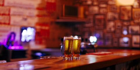 two glasses of cold beer against the background of a blurry empty beer house interior - obrazy, fototapety, plakaty