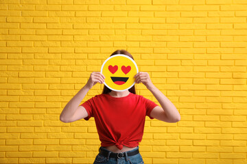 Woman covering face with heart eyes emoji near yellow brick wall
