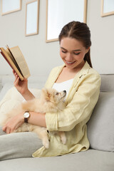 Happy young woman reading book with cute dog on sofa in living room