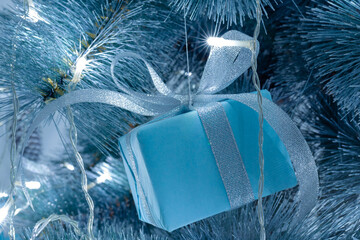 Small blue gift tied with a bow hanging on a christmas tree with blurry lights