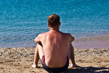 a man with a naked torso sits on the sand by the sea