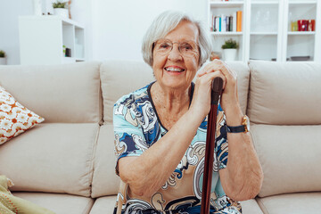 Portrait of happy mature woman in eyeglasses holding cane while sitting on sofa at home. Cheerful...