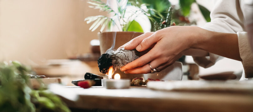 Woman hands burning white sage, palo santo before ritual on the table with candles and green plants. Smoke of smudging treats pain and stress, clear negative energy