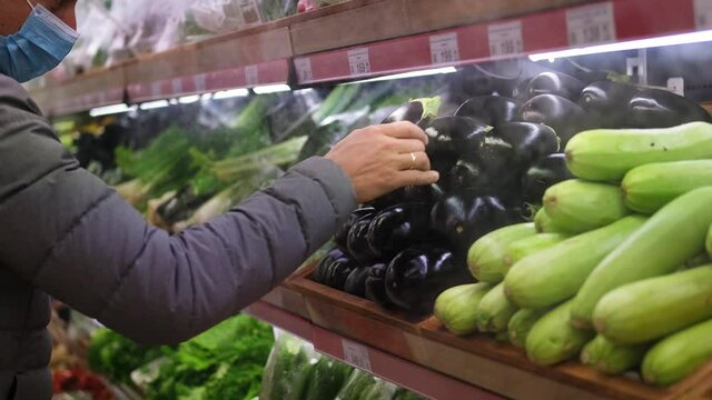 A man in a medical mask takes an eggplant from the display case of the refrigerator. Healthy eating. vegetarian. Grocery shopping amid the covid-19 pandemic.
