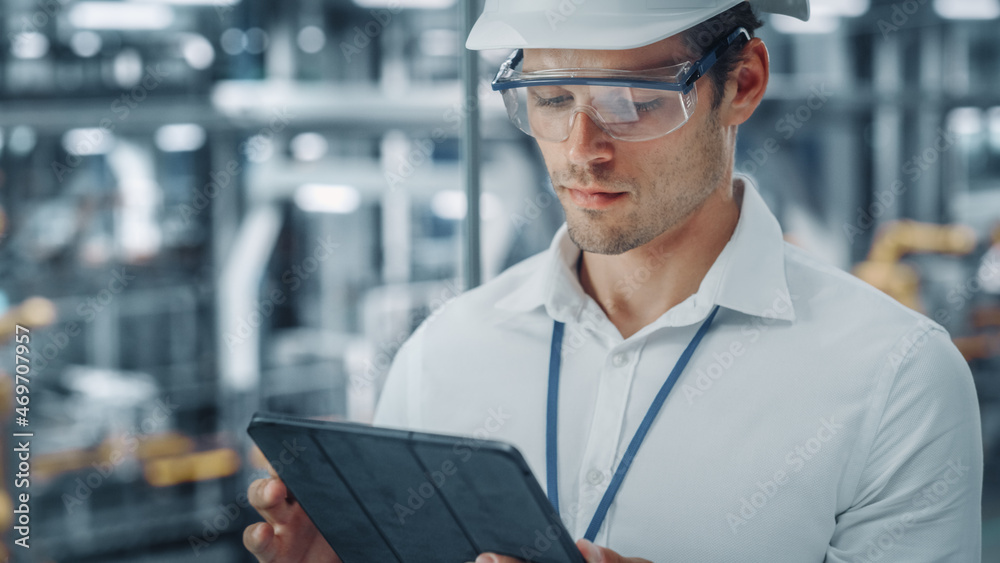 Poster male engineer in safety goggles and hard hat using tablet computer and looking out of the office at 