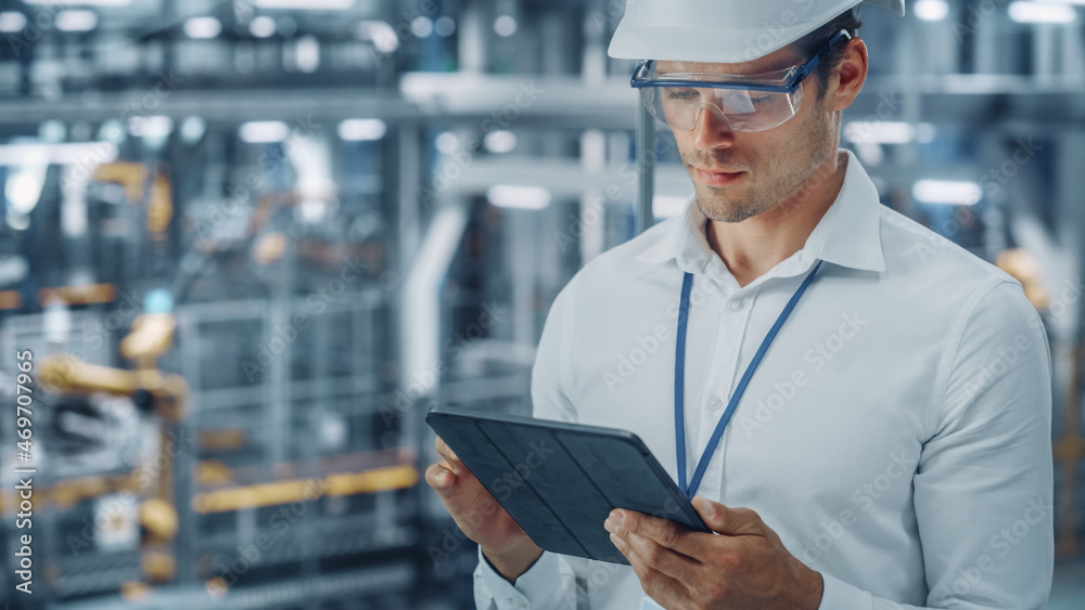 Poster male engineer in safety goggles and hard hat using tablet computer and looking out of the office at 