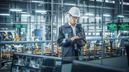 Handsome Engineer in Uniform and Hard Hat Using Tablet Computer at a Car Assembly Plant. Industrial Specialist Working on Vehicle Design, Overlooking Production in Technological Facility.