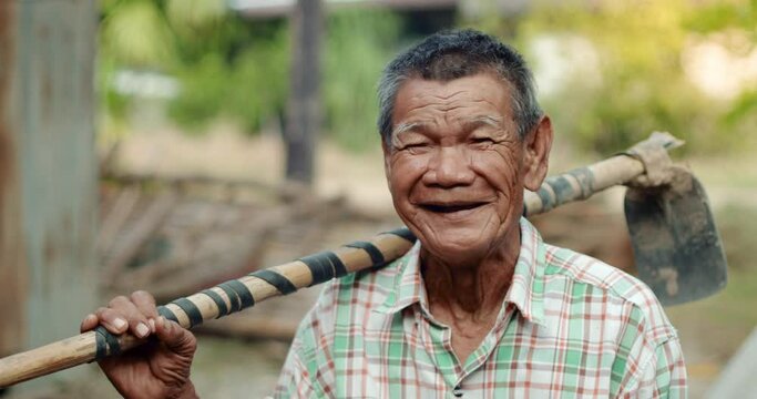 An Asian man who is more than seventy years old and works as a wage earner in a farm, carry a hoe on his shoulder, smiling and laughing happily.