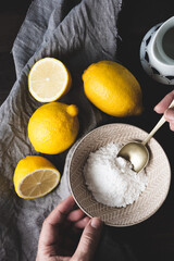 vista superior de varios limones, unas manos de mujer sosteniendo un cuenco con azúcar glas con una cuchara plateada dentro, un trapo de lino gris y una jarra sobre una mesa de madera oscura