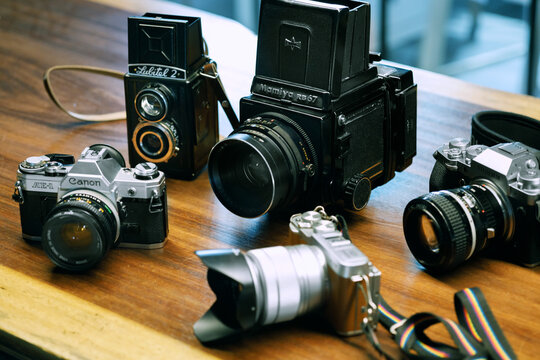 Illustrative editorial shot of Lubitell 2, Mamiya RB67, Fujifilm xt4, Canon AE1 and Fujifilm xA2 Camera on a wooden ground.