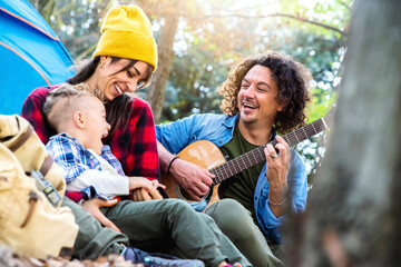 Happy family camping in the forest playing guitar and singing together - Mother, father and son having fun trekking in the nature sitting in front of the tent - Family, nature and trekking concept.