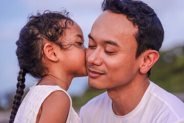 holidays, children and asian family concept -portrait of a little girl kissing her dad on cheek. Pretty girl giving a kiss to her father outdoor