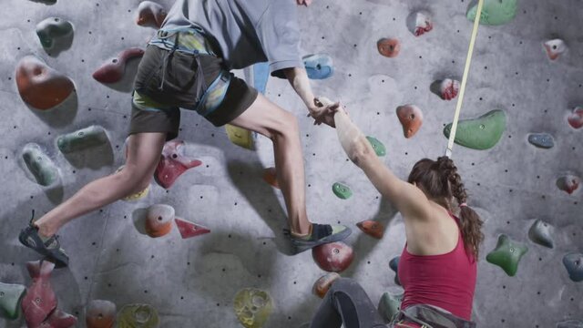 Sport girl and athletic man climb the climbing wall in the gym. The man gives a helping hand, helps to rise. Uses a safety rope on the bouldering wall. Grab hold of stones. Success and victory.