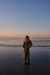 Fototapeta premium Stylish girl walks at sunset along the beach of the sea or ocean on a cool day. Travel concept. Walk on the sea or ocean. Girl in travel. Amazing scenic outdoors view. Beautiful sunset on the seashore