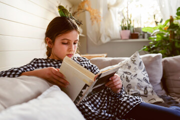 Schoolgirl reading a book at home in a living room in the company of her pet parakeet. Reading...