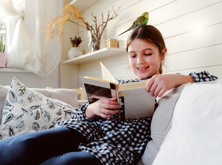 Schoolgirl reading a book at home in a living room in the company of her pet parakeet. Reading...