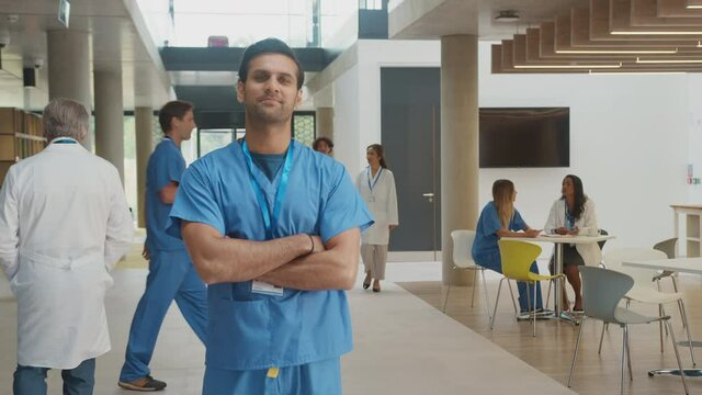 Portrait of smiling male doctor wearing scrubs standing and folding arms in busy hospital building - shot in slow motion
