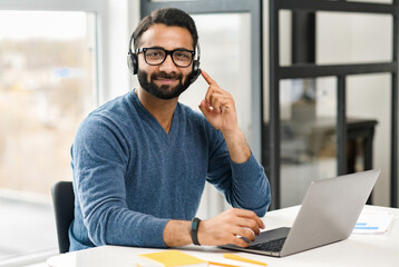Helpful and smiling Indian male employee wearing headset and eyeglasses making online consultation,...
