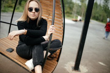 Stylish young blonde is sitting in a round hanging chair outdoor. The model sits in a chair and relaxes, looks at the camera