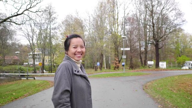 Happy Asian woman at park, smiling and looking at camera. Turn around while walking. Autumn season, leaves falling from the trees , beautiful nature, getting some fresh air at the park, Sweden
