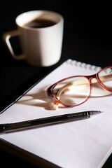 A notebook, a white teacup, glasses and a pen on a black background. Close up, top view. Office picture