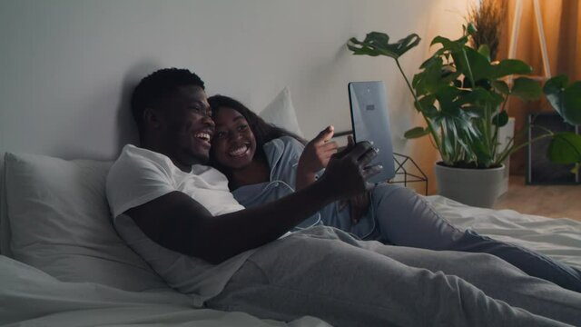 Happy african american couple wearing sleepwear embracing in bed and watching tv online on digital tablet together