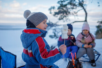 Happy friends sit around campfire on the shore of frozen lake in forest and fry marshmallows during a winter trip at weekend. Family winter picnic