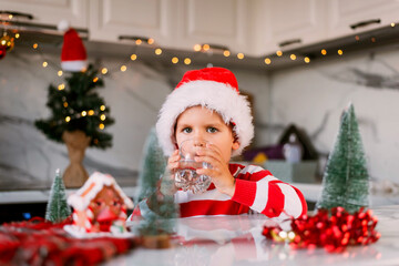 Fototapeta na wymiar Boy toddler in a red Santa hat drinking filtered water from a glass in the kitchen. Holidays, health concept.