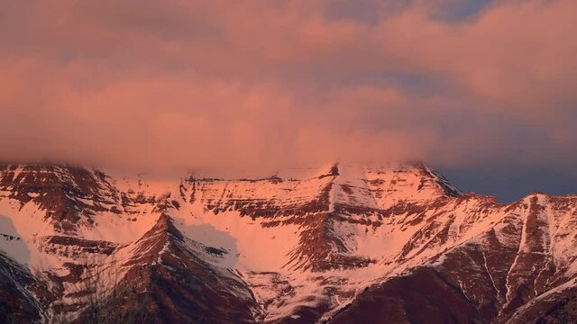 Snow capped Timpanogos Mountain during sunset with clouds moving over the peaks.