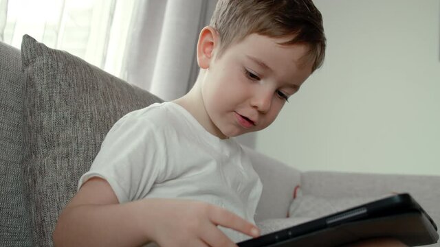 Curious cute preschool kid boy using digital tablet technology device lying on carpet floor alone. Small child hold pad computer surfing internet play game at home. Children tech addiction concept