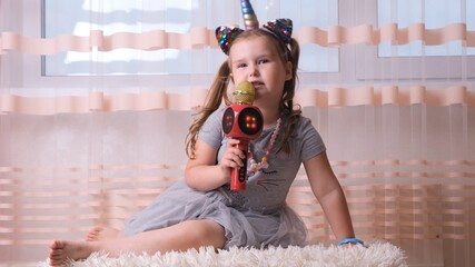 funny cute little girl 4-5 years old, singing into a karaoke microphone, with a unicorn headband,...