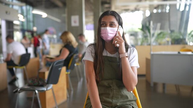 Young woman in pink protective medical face mask talking on mobile phone at indoor.
