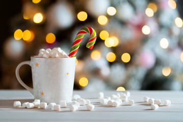 A mug of hot chocolate with marshmallow and candy stick against the background of the lights of the Christmas tree.