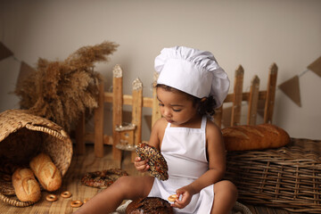 a cute dark-skinned baby with curly hair in a chef's costume has a bagel and a roll. High-quality photography